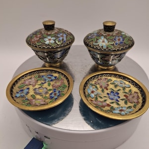 Puede incluir: Dos pequeños cuencos antiguos de cloisonné con tapas y platillos a juego. Los cuencos y platillos presentan un diseño floral en azul, rosa y verde sobre un fondo dorado. Las tapas tienen un pequeño pomo dorado.