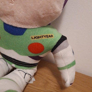 Może przedstawiać: Zielona, biała, fioletowa i czarna pluszowa zabawka Buzz Lightyear z serii Toy Story. Zabawka ma czerwone k&oacute;łko na klatce piersiowej, a na ramieniu nadrukowane jest słowo "LIGHTYEAR".