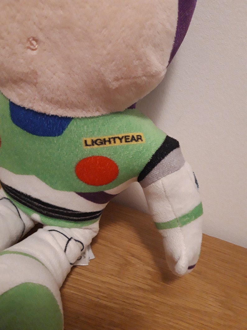 Może przedstawiać: Zielona, biała, fioletowa i czarna pluszowa zabawka Buzz Lightyear z serii Toy Story. Zabawka ma czerwone k&oacute;łko na klatce piersiowej, a na ramieniu nadrukowane jest słowo "LIGHTYEAR".