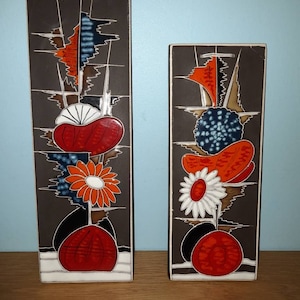 May include: Two ceramic wall plaques with abstract floral designs. The plaques are brown with white, orange, red, and blue accents. The designs feature stylized flowers, leaves, and geometric shapes.