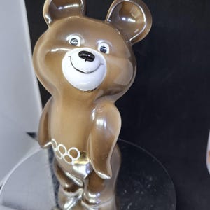 May include: A brown ceramic bear figurine with a smiling face and white accents. The bear has a gold-colored belt with the Olympic rings. The figurine is standing on a reflective surface.