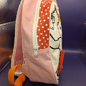 May include: A pink and white children's backpack with a cartoon horse illustration. The bag has a red polka dot pattern, a zip closure, and orange straps. The side features a small pocket. Measurements are in centimetres.