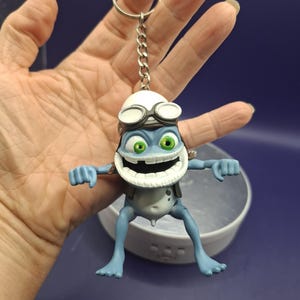 Könnte beinhalten: Ein Schlüsselanhänger in Form einer blauen, froschartigen Figur mit breitem Grinsen, großen grünen Augen und einer weißen Schutzbrille auf dem Kopf. Die Figur ist an einer silberfarbenen Metallkette mit einem runden Schlüsselring befestigt.