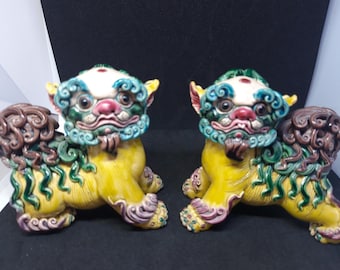 May include: Two ceramic Foo Dog statues. Each dog is yellow with green, blue, and brown accents. The dogs have detailed features, including curly manes, expressive faces, and ornate paws. They are posed in a seated position.