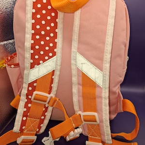 May include: Pink backpack with orange straps and white detailing. The back has a red polka dot pattern and reflective strips. Adjustable straps and a top handle are visible. A small side pocket is also present.