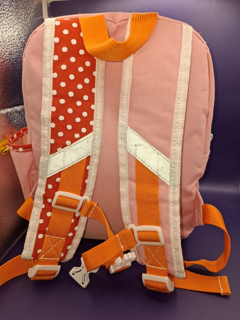 May include: Pink backpack with orange straps and white detailing. The back has a red polka dot pattern and reflective strips. Adjustable straps and a top handle are visible. A small side pocket is also present.