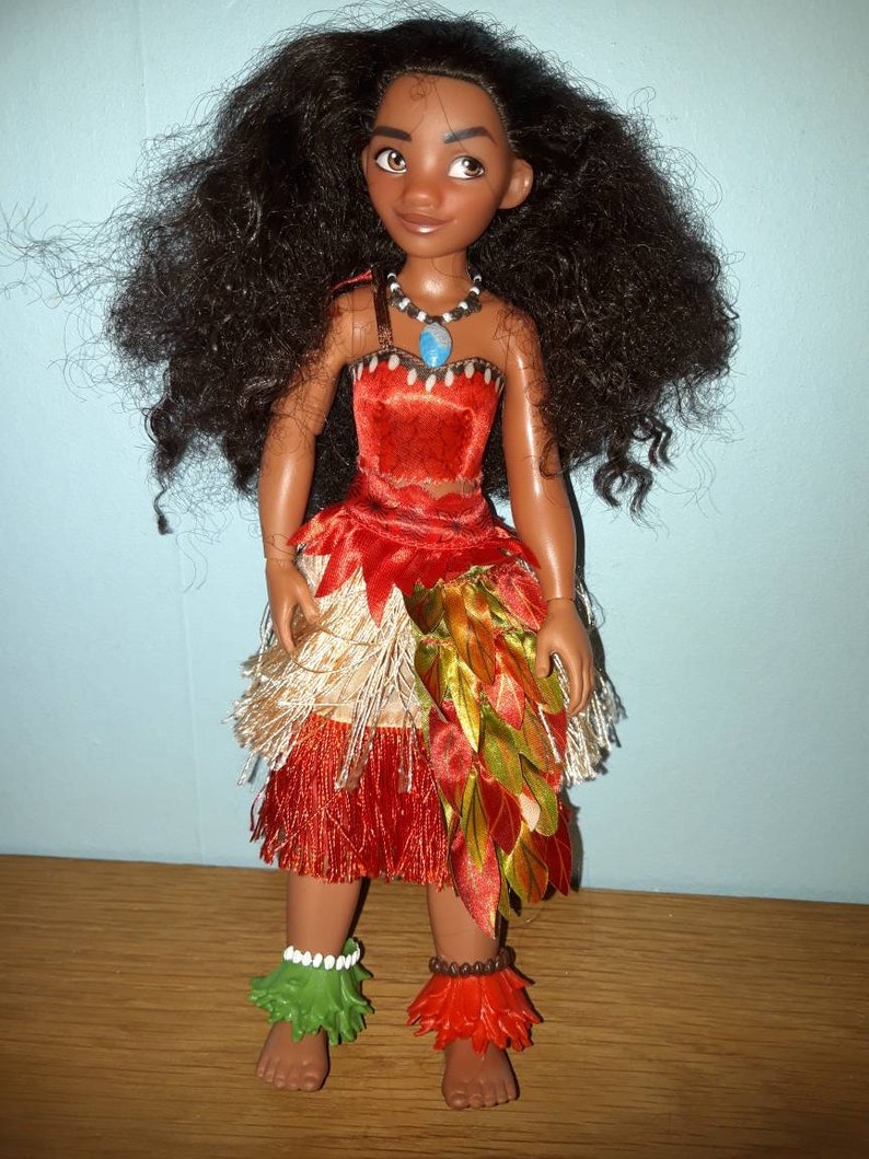 K&ouml;nnte beinhalten: Eine Moana-Puppe mit langen, lockigen braunen Haaren, die ein rotes, orangefarbenes und gr&uuml;nes Kleid mit einer blauen Halskette tr&auml;gt. Die Puppe steht auf einer Holzoberfl&auml;che.