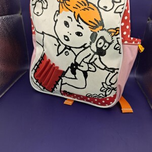 May include: White backpack featuring a cartoon illustration of a girl with orange hair and a black and white animal. The bag has red polka dot and pink accents, with orange straps and a red pocket.