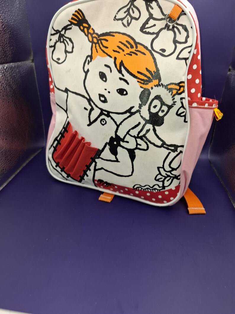 May include: White backpack featuring a cartoon illustration of a girl with orange hair and a black and white animal. The bag has red polka dot and pink accents, with orange straps and a red pocket.