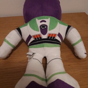 Może przedstawiać: Pluszowa zabawka Buzz Lightyear z serii Toy Story. Zabawka leży na plecach z wyciągniętymi rękami i nogami. Zabawka jest biała z zielonymi, fioletowymi i czarnymi detalami. Zabawka ma dużą głowę z fioletowym hełmem i zieloną część klatki piersiowej z czarno-białym wzorem gwiazdy.