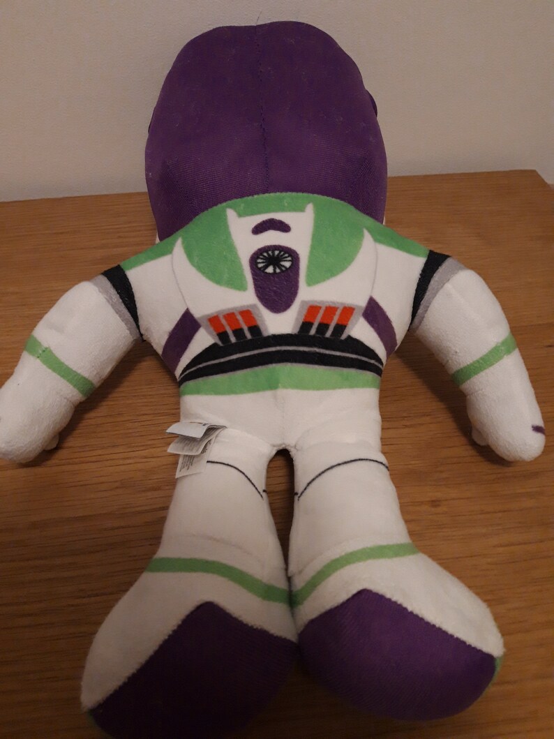 Może przedstawiać: Pluszowa zabawka Buzz Lightyear z serii Toy Story. Zabawka leży na plecach z wyciągniętymi rękami i nogami. Zabawka jest biała z zielonymi, fioletowymi i czarnymi detalami. Zabawka ma dużą głowę z fioletowym hełmem i zieloną część klatki piersiowej z czarno-białym wzorem gwiazdy.