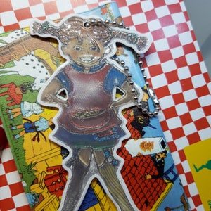 Pu&ograve; includere: Un portachiavi in ​​argento con un ciondolo a forma di ragazza dei cartoni animati con le trecce che indossa un vestito blu e rosso. Il ciondolo &egrave; attaccato a una catena d'argento.