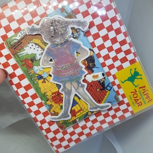 Pu&ograve; includere: Uno sfondo a scacchi rosso e bianco con un coperchio in plastica trasparente. Un'illustrazione colorata di Pippi Calzelunghe &egrave; attaccata allo sfondo. L'illustrazione &egrave; un ritaglio di Pippi Calzelunghe in piedi con le braccia sui fianchi. L'illustrazione &egrave; circondata da un bordo bianco. Il testo "Pippi Langstrump 70 &aring;r 1945-2015" &egrave; stampato su un rettangolo giallo nell'angolo in basso a destra dell'immagine.