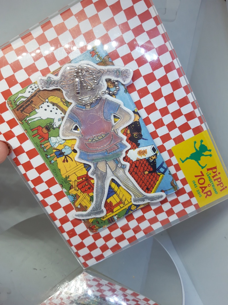 Pu&ograve; includere: Uno sfondo a scacchi rosso e bianco con un coperchio in plastica trasparente. Un'illustrazione colorata di Pippi Calzelunghe &egrave; attaccata allo sfondo. L'illustrazione &egrave; un ritaglio di Pippi Calzelunghe in piedi con le braccia sui fianchi. L'illustrazione &egrave; circondata da un bordo bianco. Il testo "Pippi Langstrump 70 &aring;r 1945-2015" &egrave; stampato su un rettangolo giallo nell'angolo in basso a destra dell'immagine.