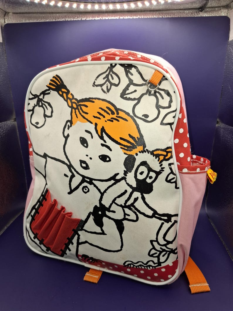 May include: A backpack featuring a cartoon illustration of a girl with orange hair and a monkey. The backpack has a white front with red polka dots, pink sides, and orange straps. The illustration is outlined in black.