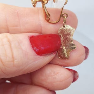 May include: Gold-tone clip-on earrings with a dangling charm featuring a cartoon character. The charm is inscribed with the word "Disney".