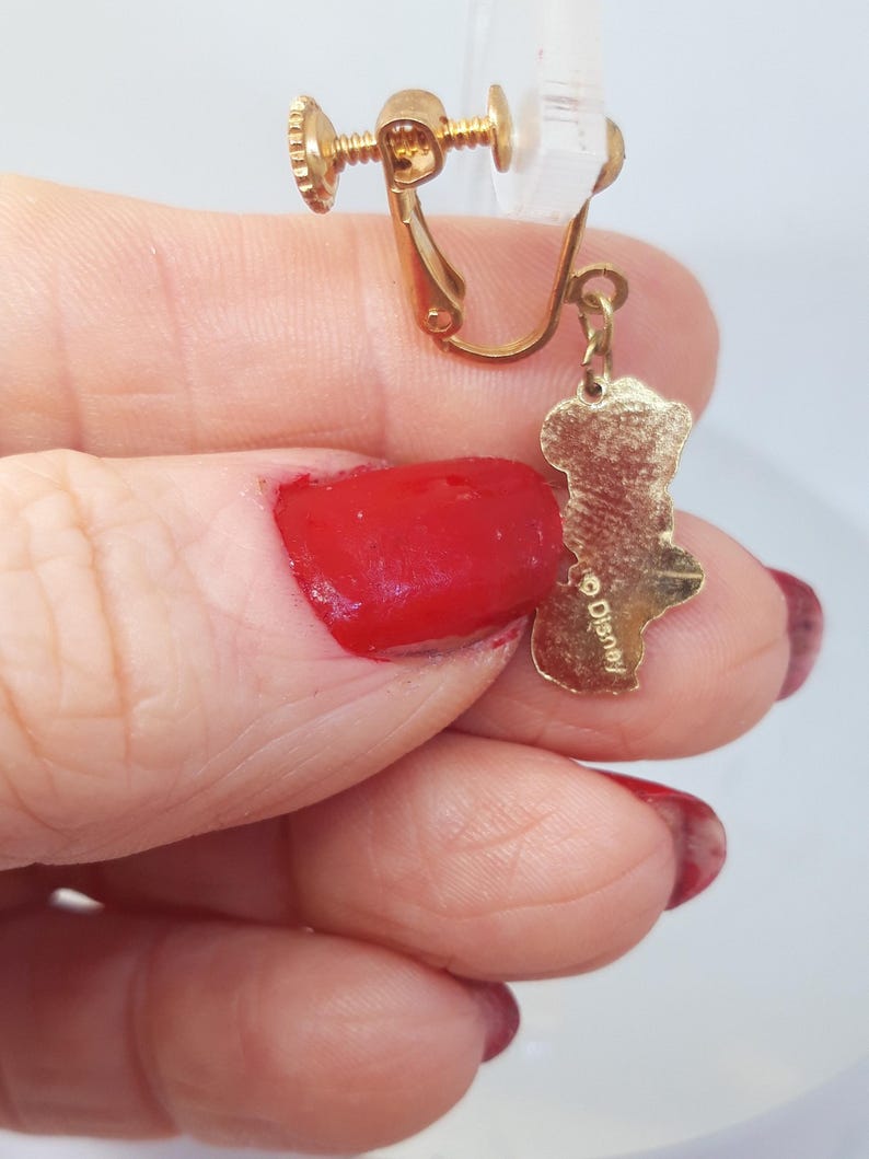 May include: Gold-tone clip-on earrings with a dangling charm featuring a cartoon character. The charm is inscribed with the word "Disney".