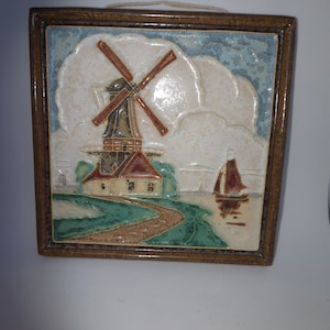 May include: A framed ceramic tile featuring a raised image of a windmill and a small house in a landscape with a sailboat in the distance. The tile is surrounded by a brown wooden frame.