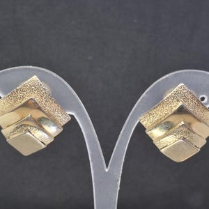 May include: Gold-toned square earrings with a layered, geometric design. The earrings have a textured, granular finish on the top layer and a smooth finish on the lower layers. The earrings are displayed on a clear earring stand against a dark background.