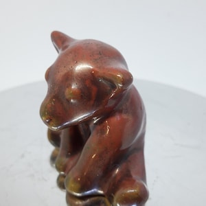 Può includere: Una piccola statuetta in ceramica di un orso bruno seduto sulle zampe posteriori. L'orso ha una finitura lucida ed è dipinto in tonalità di marrone e rosso.