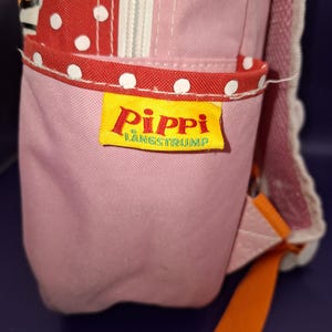May include: Pink backpack with red polka dot detailing and a yellow label reading "Pippi L&aring;ngstrump". The bag features an orange strap and a white zip. The bag is made of a durable fabric.