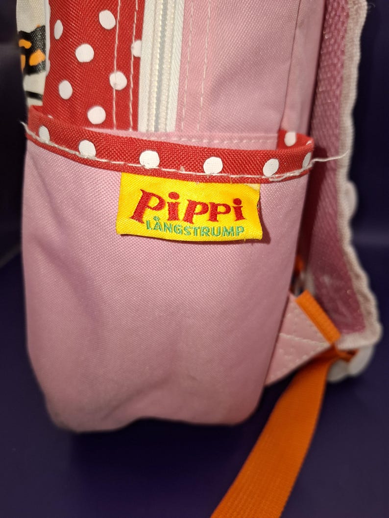 May include: Pink backpack with red polka dot detailing and a yellow label reading "Pippi L&aring;ngstrump". The bag features an orange strap and a white zip. The bag is made of a durable fabric.