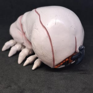 Puede incluir: Una escultura blanca y rosa con forma de insecto con múltiples patas y un cuerpo redondeado. La escultura tiene un apéndice azul oscuro y detalles naranjas y negros en la parte inferior. El objeto está sobre un fondo oscuro.