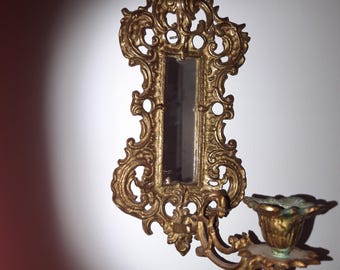 May include: A gold-toned wall sconce with a small mirror and a candle holder. The sconce has a decorative, ornate design.