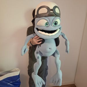 May include: A large, light blue plush character figure featuring a wide, toothy grin, prominent green eyes, and a white helmet with dark brown goggles. The character has long, thin limbs with four-fingered hands and feet, and dark blue spots.