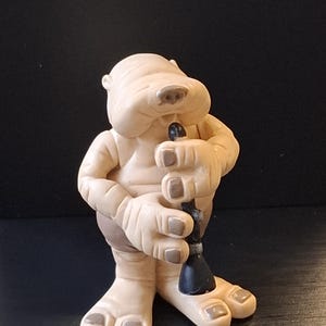 Può includere: Statuetta beige di una creatura simile a un cartone animato che tiene in mano un oggetto nero. La figura ha mani e piedi grandi con accenti marroni. Lo sfondo è nero.