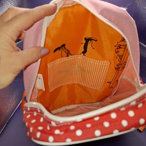 May include: Pink backpack with a red polka dot base and white trim. The interior is orange with black cartoon figures and a white striped pocket. The backpack is open, showing the inside.