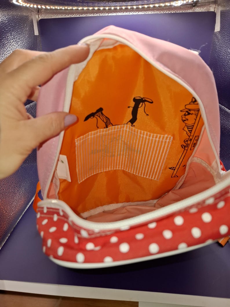 May include: Pink backpack with a red polka dot base and white trim. The interior is orange with black cartoon figures and a white striped pocket. The backpack is open, showing the inside.