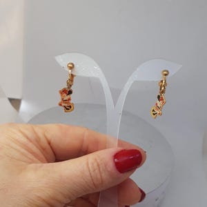 May include: A pair of gold-tone clip-on earrings featuring a cartoon character with red, yellow, and black details. The earrings are displayed on a white stand.