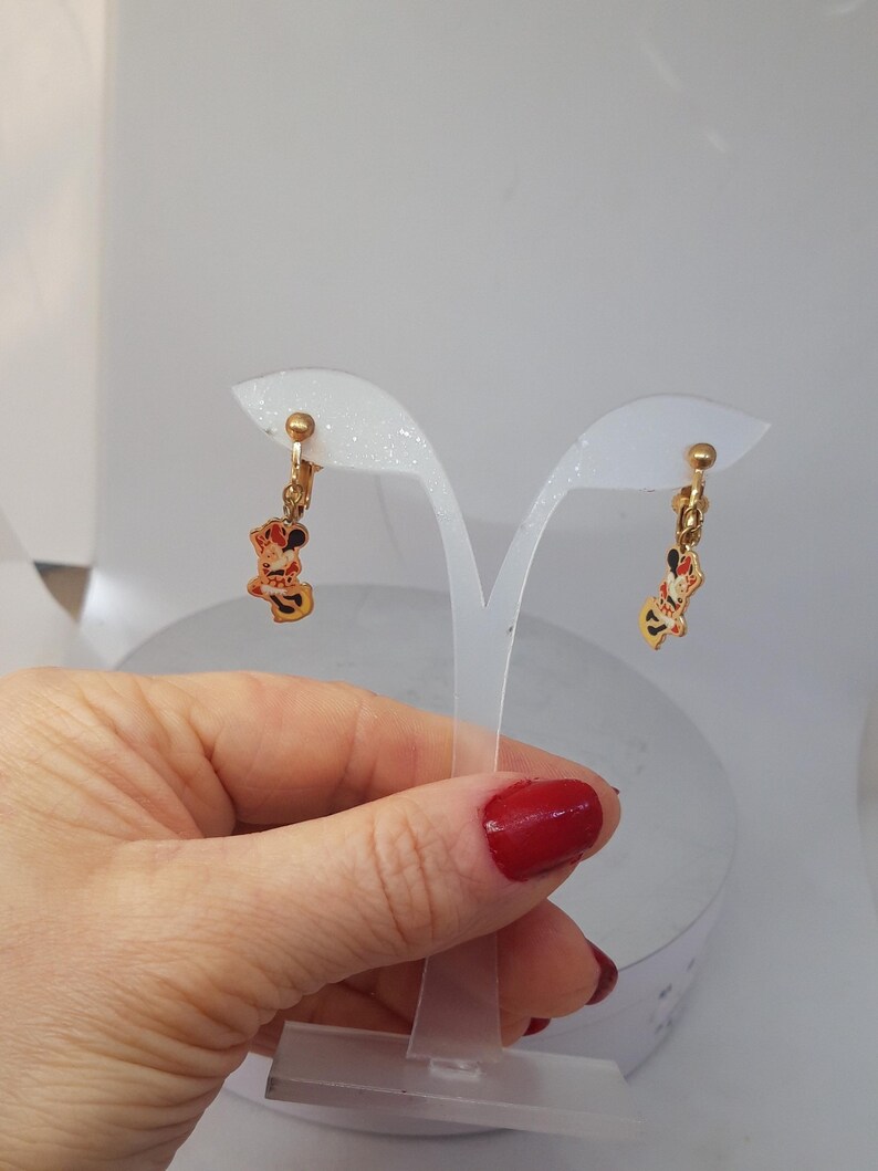 May include: A pair of gold-tone clip-on earrings featuring a cartoon character with red, yellow, and black details. The earrings are displayed on a white stand.