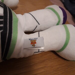 Może przedstawiać: Biała pluszowa zabawka Buzz Lightyear z serii Toy Story. Zabawka ma zielone i fioletowe akcenty oraz czarno-biały pasiasty wz&oacute;r na klatce piersiowej. Zabawka ma metkę z napisem "Disney Pixar Toy Story 4".