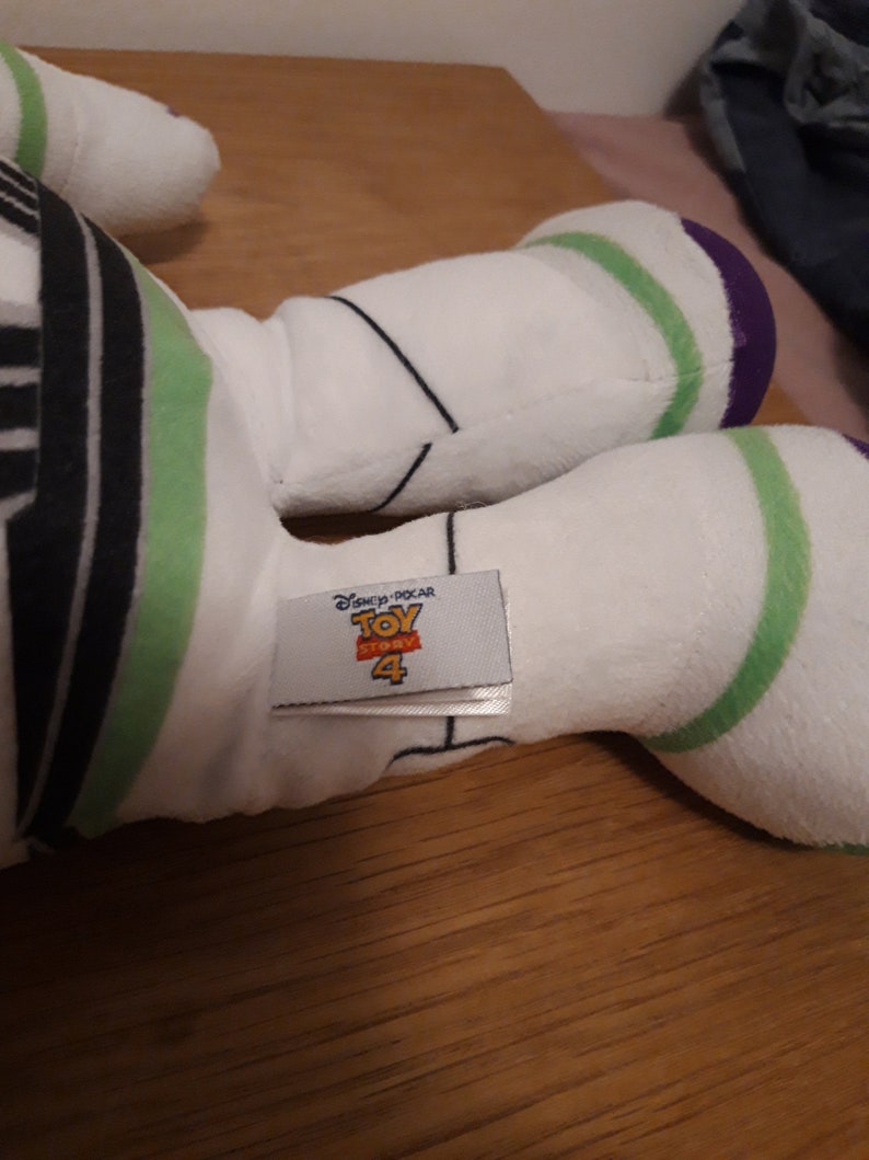 Może przedstawiać: Biała pluszowa zabawka Buzz Lightyear z serii Toy Story. Zabawka ma zielone i fioletowe akcenty oraz czarno-biały pasiasty wz&oacute;r na klatce piersiowej. Zabawka ma metkę z napisem "Disney Pixar Toy Story 4".