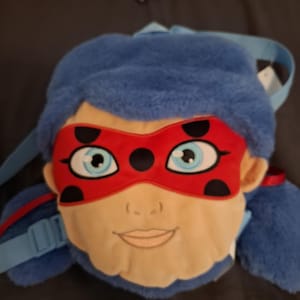 Pode incluir: Uma mochila de pelúcia com o rosto de uma personagem de desenho animado com cabelo azul, uma máscara vermelha com bolinhas pretas e olhos azul claro. A mochila tem alças azuis e um rosto bege.