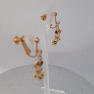 May include: A pair of gold-tone clip-on earrings with a dangling charm of a cartoon character. The charm is white with red and black details.