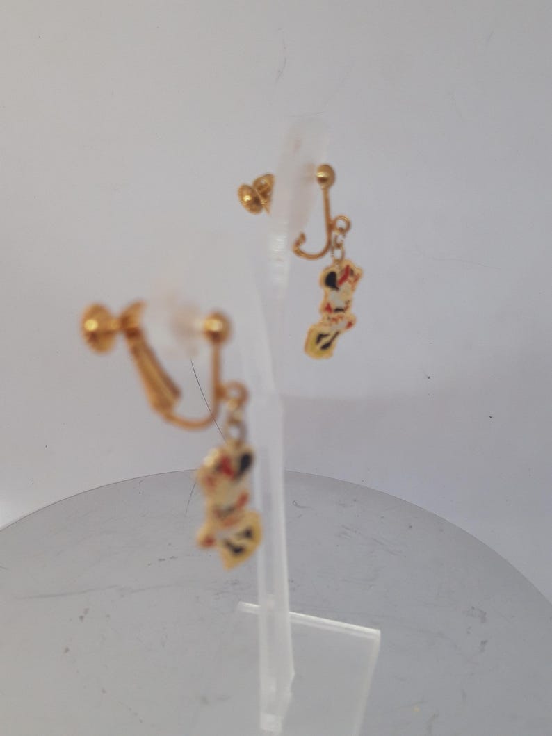 May include: A pair of gold-tone clip-on earrings with a dangling charm of a cartoon character. The charm is white with red and black details.