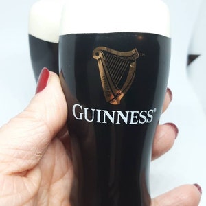 Puede incluir: Dos vasos de chupito de cer&aacute;mica negra con una parte superior de espuma blanca. Los vasos presentan el logotipo de Guinness, un arpa dorada y el texto "ESTD 1778" y "GUINNESS".