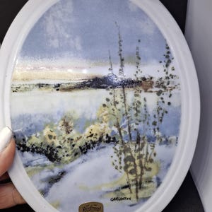 Könnte beinhalten: Ovale Porzellanteller mit einer Landschaftsszene. Der Teller hat einen weißen Rand und ein gemaltes Bild eines Sees, Bäumen und einem bewölkten Himmel. Der Teller trägt den Text "Carlanton" und ein goldenes Emblem mit der Aufschrift "Portlands Sweden".