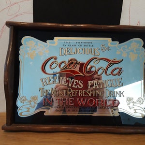 以下が含まれることがあります： 木製のフレームが付いたヴィンテージのCoca-Colaミラートレイ。ミラーは、青と金のデザインで、「どこでも販売されています。ガラスまたはボトル入り。おいしい5セント。コカ・コーラは疲労を和らげます。世界で最もさわやかな飲み物。ジンジャーエールやソーダがおいしい場所ならどこでも。」というテキストが書かれています。