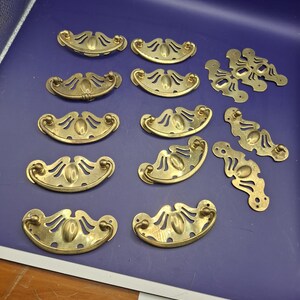 Peut inclure: Une collection de dix poignées de tiroir en laiton disposées en rangées. Chaque poignée présente un design incurvé en demi-cercle avec un centre ovale et des découpes décoratives. La quincaillerie comprend des plaques arrière en laiton assorties.