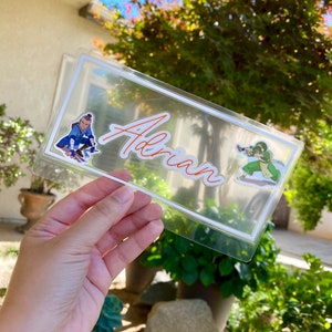 May include: A clear plastic frame with a white border, featuring the name "Adrian" written in orange cursive. Two cartoon characters are visible on the frame, one in blue and one in green.