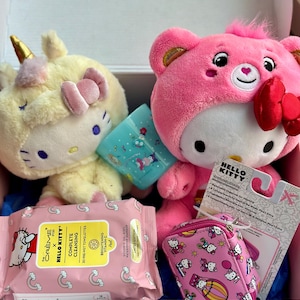 May include: A pink Hello Kitty plush toy wearing a pink bear costume with a red bow. A yellow Hello Kitty plush toy wearing a unicorn costume. A pink Hello Kitty makeup bag with a zipper. A package of Hello Kitty makeup remover wipes. The text on the package reads "CRÈME x HELLO KITTY COMPLETE CLEANSING 30 PRE-WET TOWELETTES BRIGHTENING VITAMIN C Peel GOODBYE OIL, DIRT, WATERPROOF MAKEUP LIMITED EDITION".