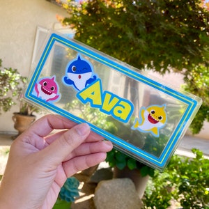 May include: A clear plastic sign with a blue border and yellow stripes. The sign has a blue and white shark, a pink and white shark, and the name "Ava" in yellow letters.