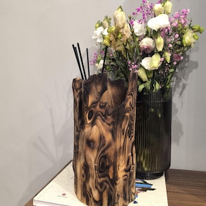 May include: A rustic, cylindrical wooden vase with a dark, textured finish, holding black reeds. Beside it, a glass vase displays a bouquet of white, pink, and yellow flowers. The vase sits on a book.