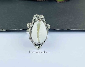 May include: A silver ring with a white cowrie shell set in a decorative filigree design. The ring is on a white surface with the text "krittikajewelers" below it.