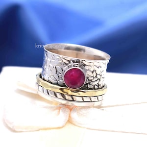 May include: A silver and gold spinning ring with a red gemstone. The ring has a textured band with a floral design.