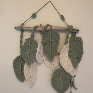 May include: A wall hanging made of macrame with green and white feathers hanging from a driftwood branch.